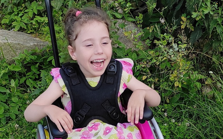 Uśmiechnięta dziewczynka w wózku, otoczona zielenią.