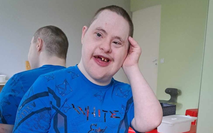 Chłopiec z uśmiechem, siedzący w pokoju, w niebieskiej koszulce.