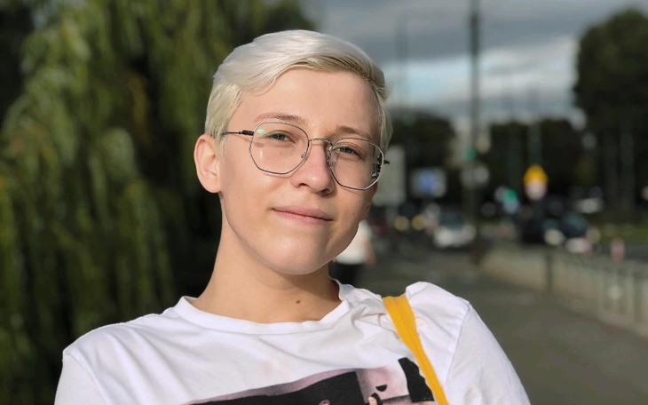 Młody człowiek z okularami, uśmiechnięty, na tle miejskiego krajobrazu.