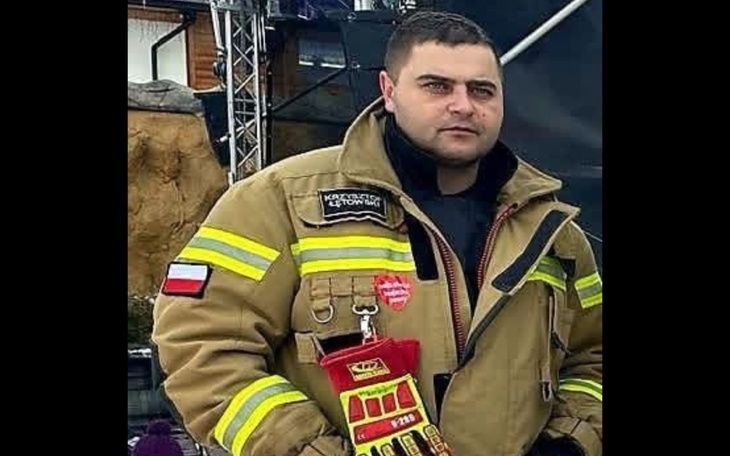 Mężczyzna w stroju strażaka na tle sceny, czerwone akcenty na ubraniu i sprzęt