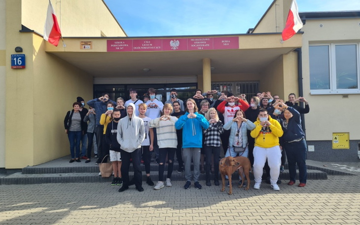 Grupa ludzi przed budynkiem, wszyscy w pozycji z sercami.