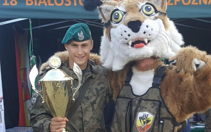 Mężczyzna w mundurze z pucharem, obok maskotka w kształcie rysia.