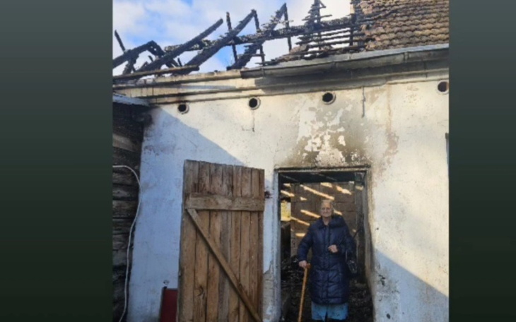 Starsza kobieta stojąca przed zniszczonym domem.