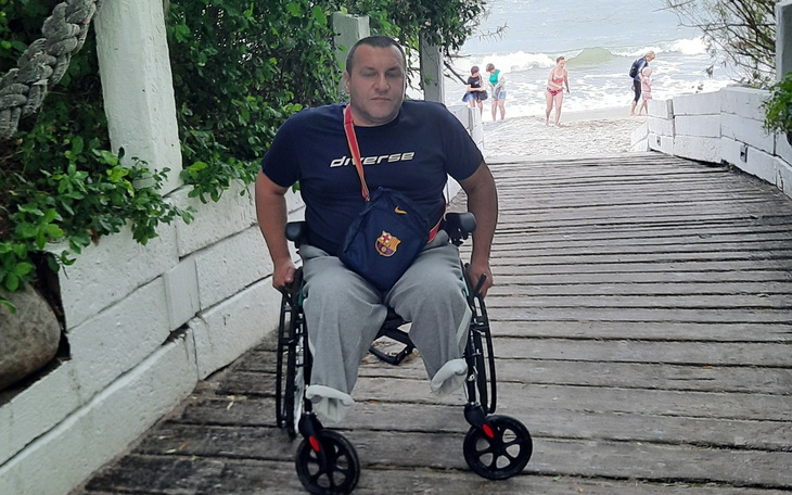 Mężczyzna na wózku inwalidzkim na plaży, w tle ludzie.