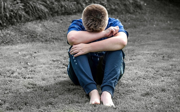 Chłopiec siedzący na trawie, zasłaniający twarz, wyrażający smutek.