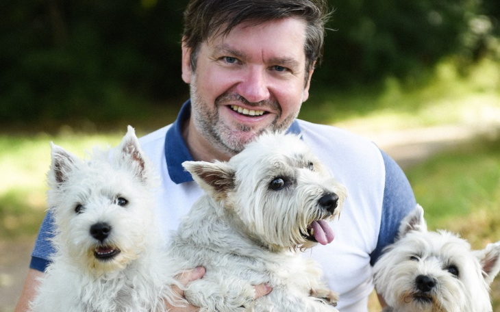 Mężczyzna z trzema psami, uśmiechnięty na tle natury.