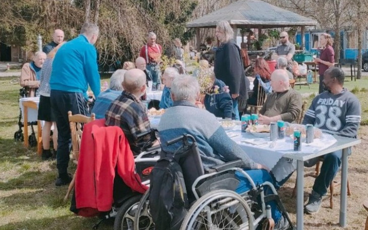 Grupa ludzi przy stole, cieszących się wspólnym posiłkiem na świeżym powietrzu.