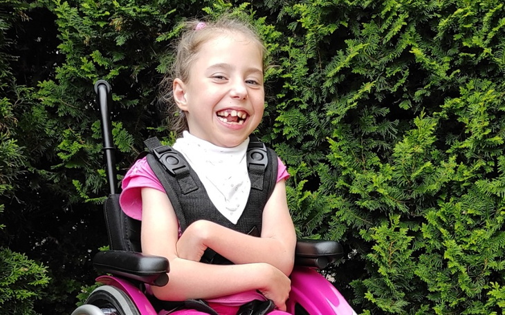 Uśmiechnięta dziewczynka w różowym wózku inwalidzkim, otoczona zielonymi krzewami.
