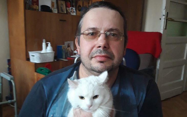 Mężczyzna trzyma białego kota na rękach, obaj patrzą w kamerę.