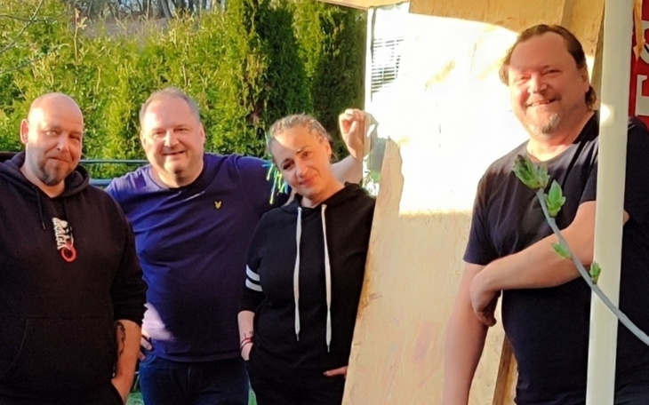 Grupa ludzi pozujących na tle drewnianej konstrukcji w słoneczny dzień.