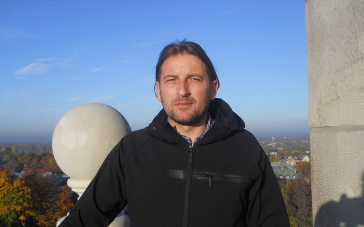 Mężczyzna na tle nieba, w czarnej kurtce, z widokiem na miasto.