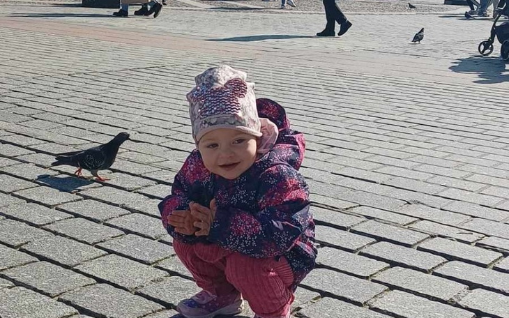 Mała dziewczynka w kolorowej kurtce bawi się na placu, obok niej ptak.