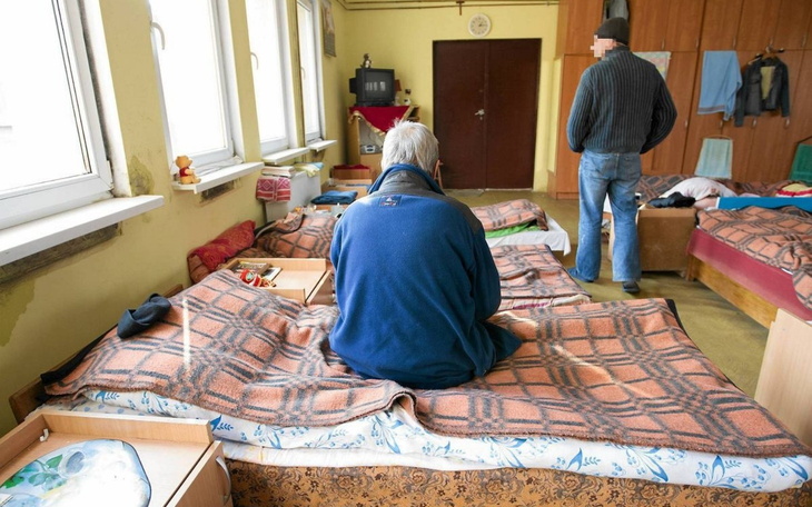 Osoba siedząca na łóżku w schronisku, z widokiem na inne łóżka.