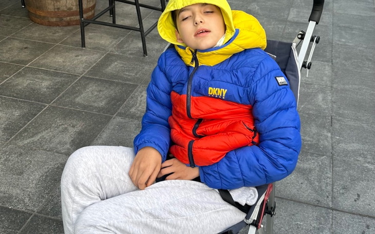 Chłopiec w kolorowej kurtce siedzący na wózku, z zamkniętymi oczami.