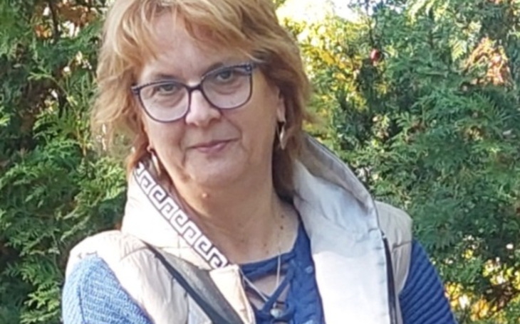 Kobieta z okularami i krótkimi włosami, uśmiechająca się w otoczeniu zieleni.