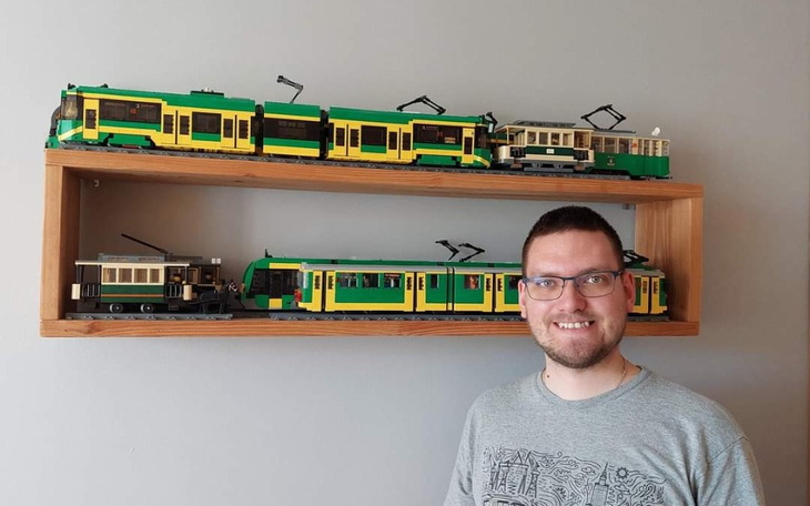 Mężczyzna z uśmiechem obok modelu tramwajów LEGO na półce.