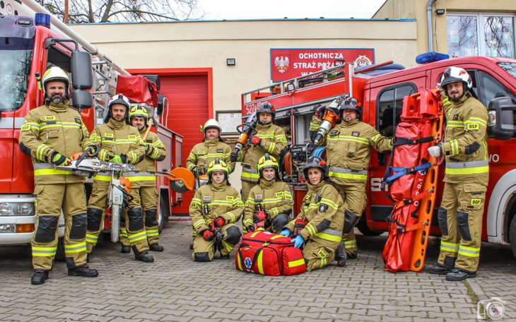 Grupa strażaków w mundurach, pozująca przed wozem strażackim.