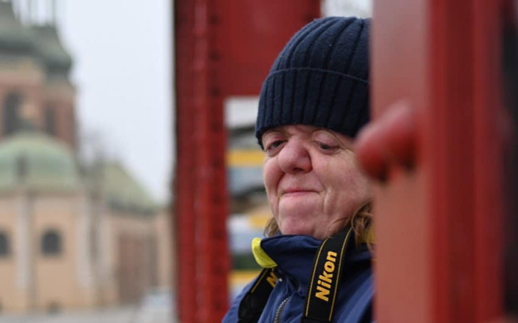 Kobieta w czapce, z uśmiechem, stojąca przy czerwonym elemencie architektury.