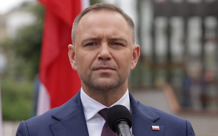 Mężczyzna w garniturze przemawia, w tle flagi.