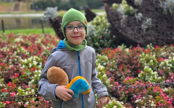 Chłopiec w zielonym czapce trzymający pluszaka wśród kolorowych kwiatów.