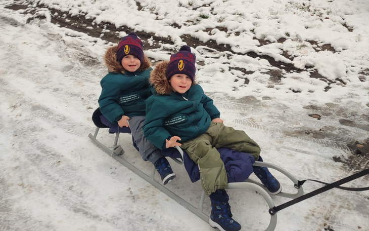 Dwoje dzieci na sankach, ubrane w ciepłe kurtki i czapki, bawią się na śniegu.