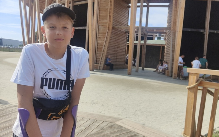 Chłopiec w koszulce Puma stoi przed drewnianą konstrukcją.