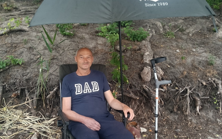 Mężczyzna siedzi w krześle pod parasolem w plenerze, uśmiecha się spokojnie.