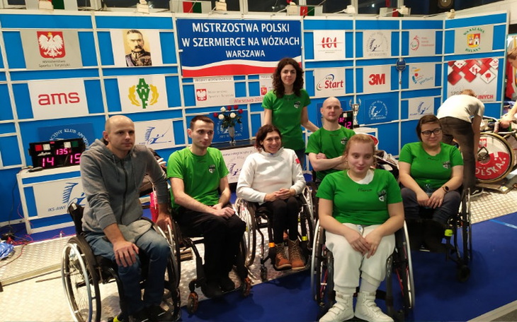 Grupa osób w zielonych koszulkach na tle planszy z logo sponsorów.