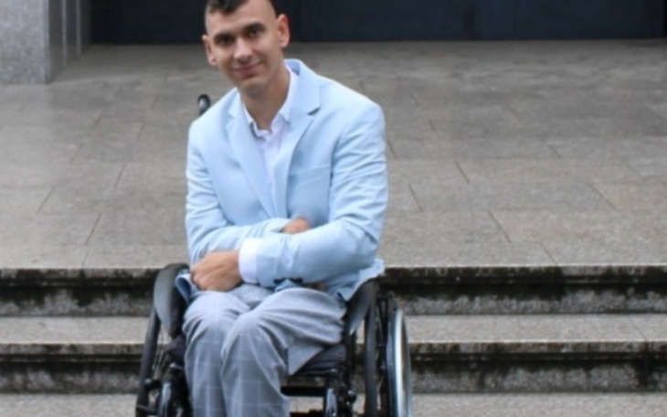 Mężczyzna w niebieskim garniturze na wózku inwalidzkim, uśmiechający się.