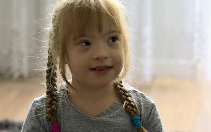 Dziewczynka z warkoczami, uśmiechająca się, siedzi w jasnym pomieszczeniu.