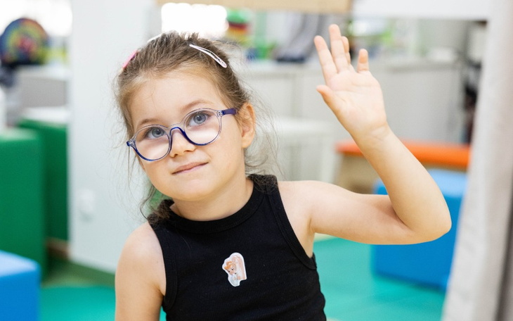 Mała dziewczynka w okularach, machająca ręką w radosny sposób.