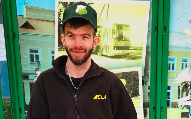 Mężczyzna w czapce z uśmiechem, stojący przed zieloną ścianą z plakatami.