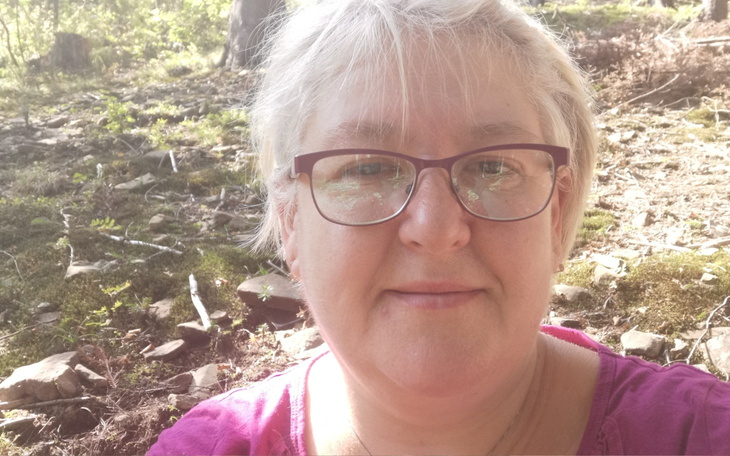 Kobieta w okularach na tle leśnej scenerii, uśmiechnięta.