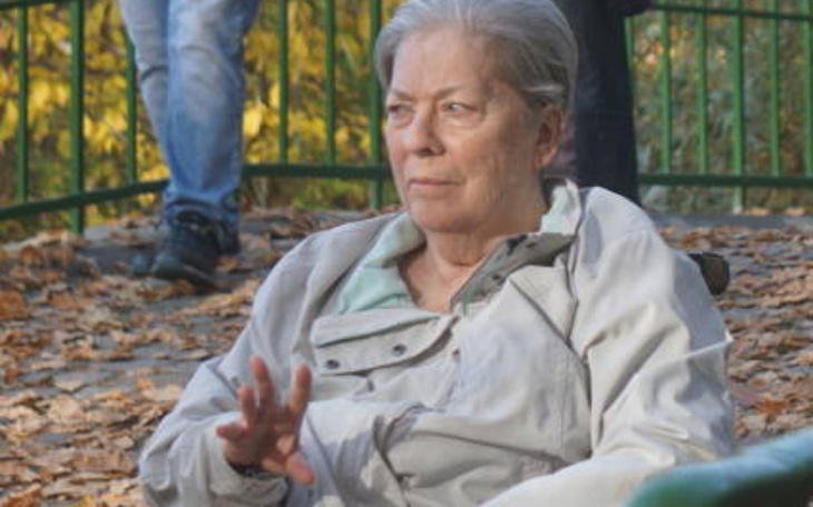 Starsza kobieta siedząca na ławce w parku, otoczona liśćmi.