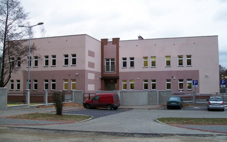 Budynek szkoły z parkingiem, w otoczeniu drzew.