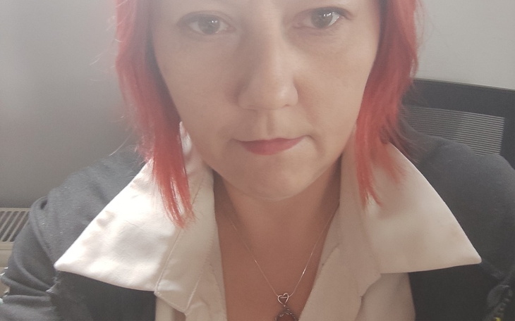 Portret kobiety z czerwonymi włosami, w białej koszuli, siedzącej przy biurku.
