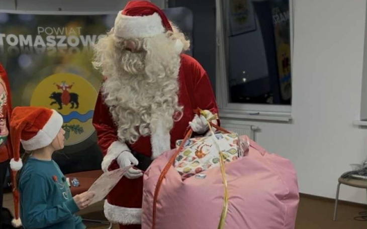 Święty Mikołaj wręcza prezent dziecku w czerwonej czapce.