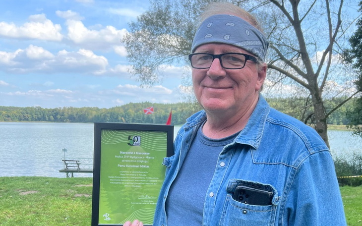 Mężczyzna trzymający dyplom nad jeziorem w słoneczny dzień.