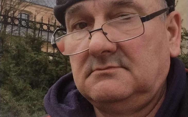 Mężczyzna w okularach, z poważnym wyrazem twarzy, na tle zieleni.