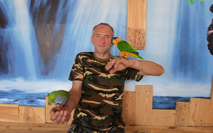 Mężczyzna w kamuflażu trzyma dwa kolorowe papugi na rękach.