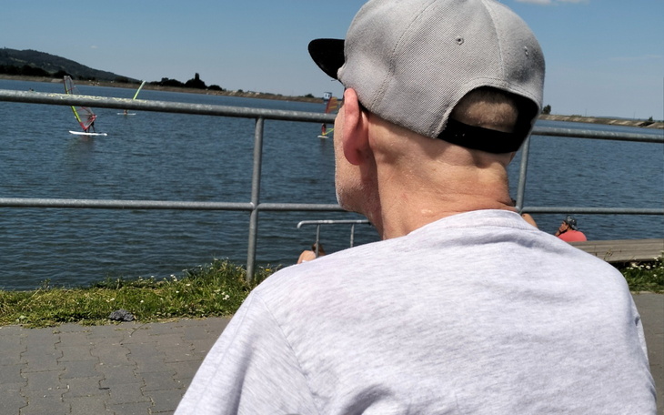 Mężczyzna siedzący nad wodą, obserwujący windsurfing.