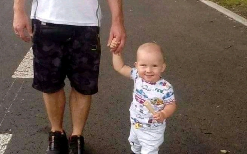 
        Małe dziecko trzyma się za rękę dorosłego, uśmiechnięte na spacerze.
    