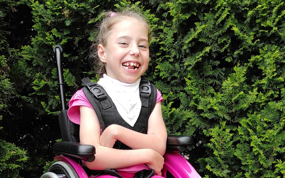 
        Uśmiechnięta dziewczynka w różowym wózku inwalidzkim, otoczona zielonymi krzewami.
    