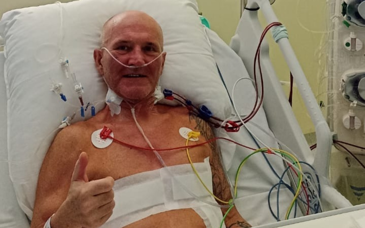 
        Mężczyzna w szpitalu, uśmiechający się z palcami w górze.
    