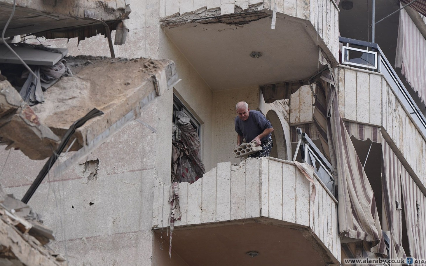 
        Mężczyzna na zniszczonym balkonie, trzymający cegłę.
    