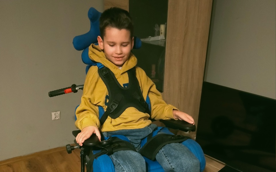 
        Chłopiec w specjalnym wózku, uśmiechający się.
    