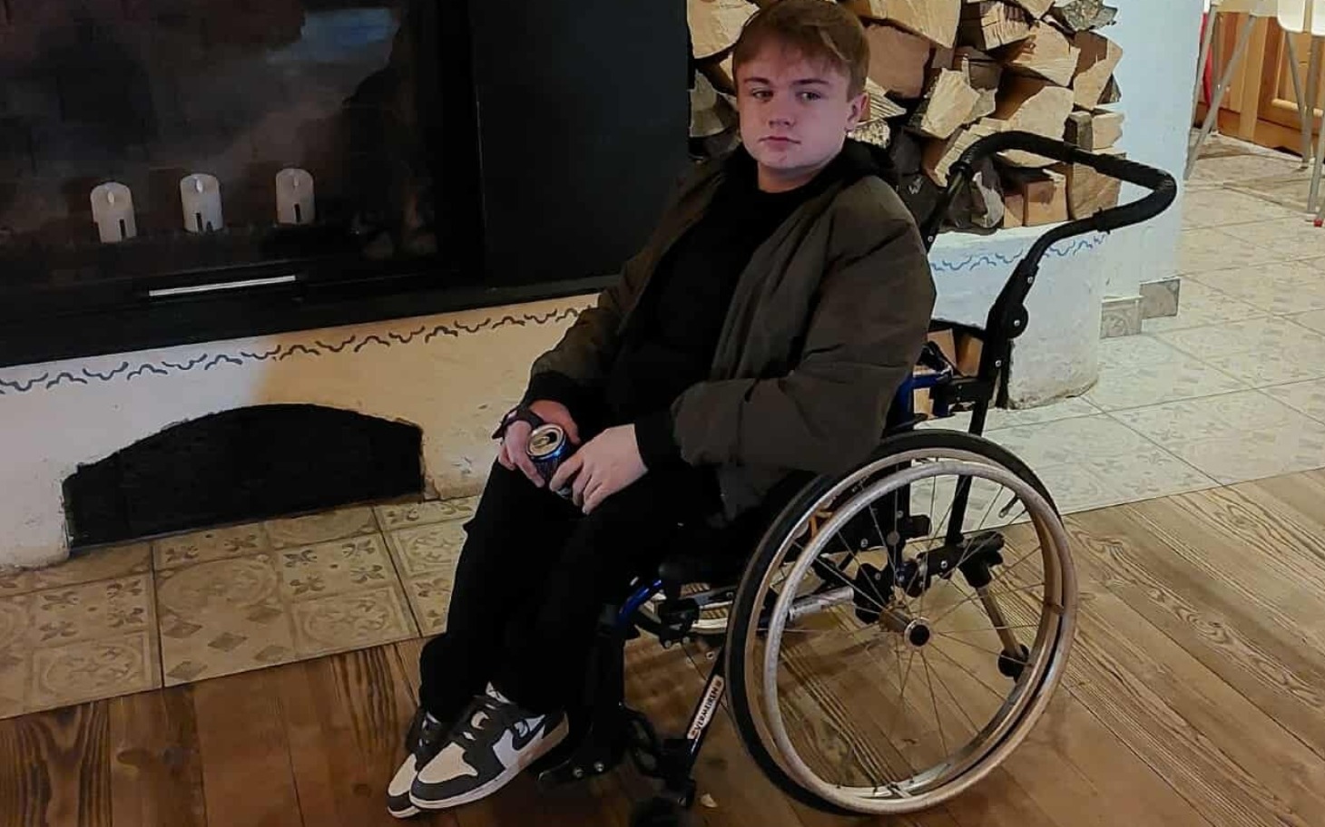 Osoba w wózku inwalidzkim, trzymająca napój, w otoczeniu drewna.