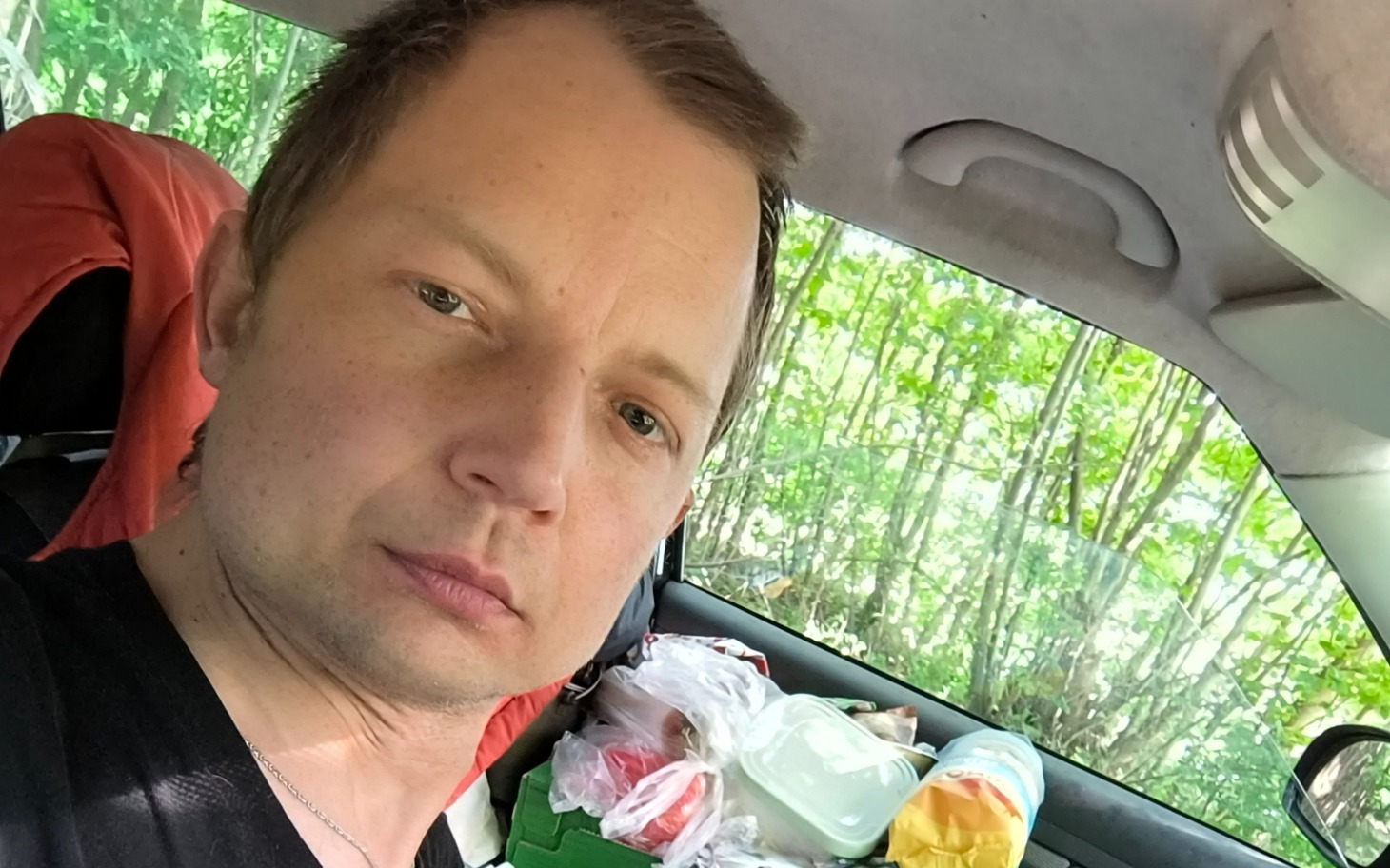 
         Człowiek w samochodzie z rzeczami w bagażniku, zieleń za szybą, samotne selfie.
    