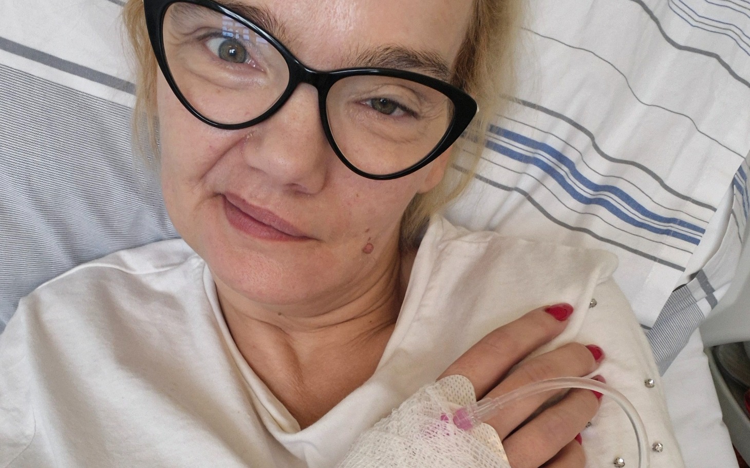 
        Kobieta w łóżku szpitalnym, z kroplówką i uśmiechem.
    