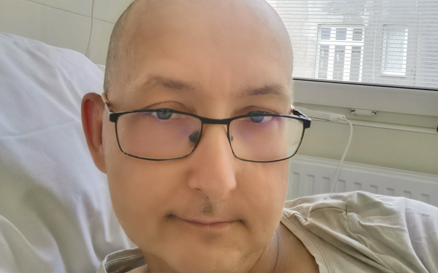 
        Mężczyzna w łóżku szpitalnym, z okularami i spokojnym wyrazem twarzy.
    